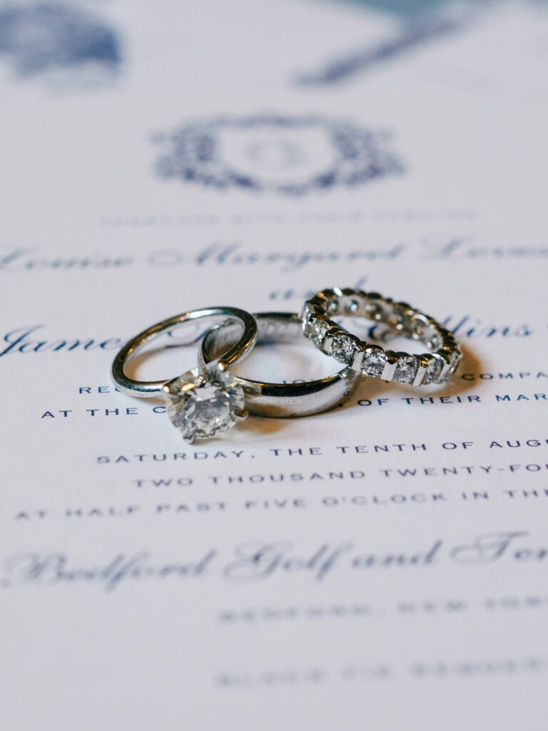 Close-up of three wedding rings resting on the invitation, showcasing a solitaire diamond engagement ring and diamond eternity band