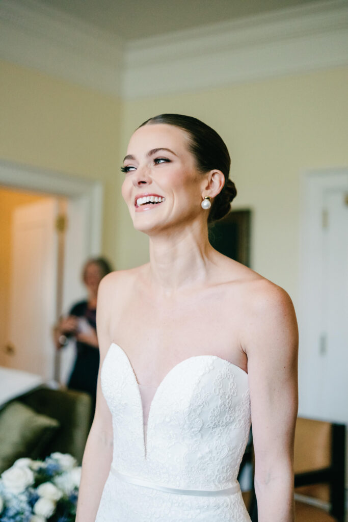Bride laughing with joy in the bridal suite, wearing a strapless lace gown and pearl earrings just moments before the first look
