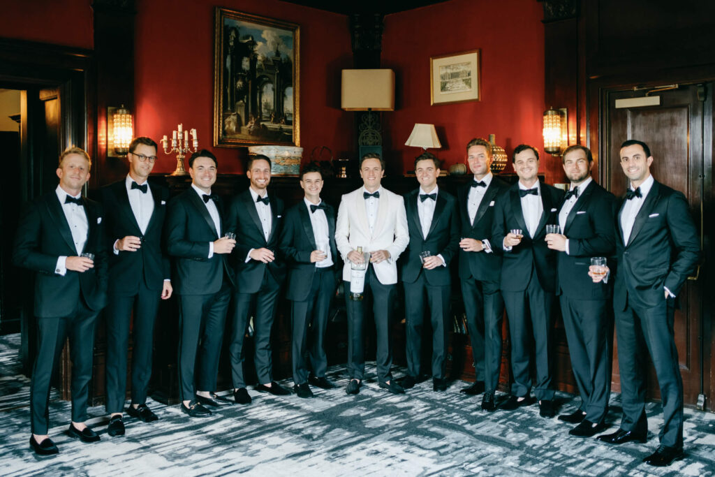 Group portrait of groom with groomsmen in classic black tuxedos inside the Castle Hotel library, raising glasses before the wedding ceremony