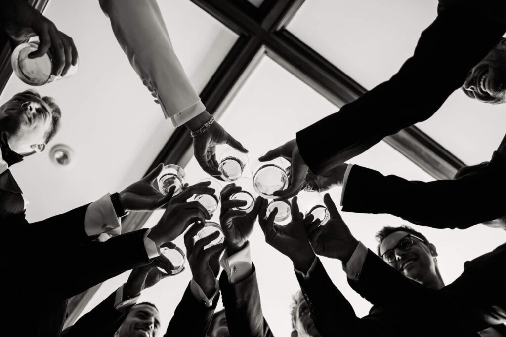 Black and white upward shot of the groom and groomsmen clinking glasses in a celebratory toast, framed by natural light from above