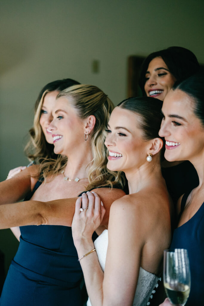 Bride taking a joyful selfie with her bridesmaids in navy gowns, all smiling and holding champagne flutes before the wedding begins