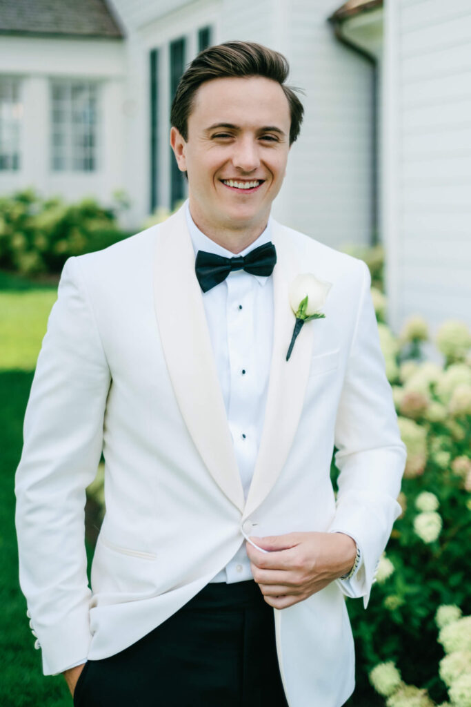 Groom smiling outdoors in front of hydrangeas, wearing a white tuxedo jacket and boutonnière before the New York country club wedding