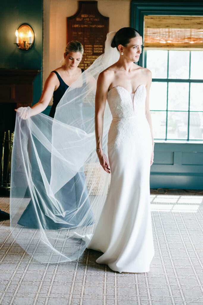 Bridesmaid helping the bride adjust her flowing veil inside Bedford Golf & Tennis Club, with natural light filtering through the windows