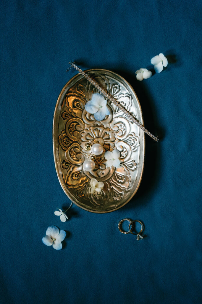 Elegant bridal jewelry flat lay with pearl earrings, diamond bracelet, and delicate blue hydrangeas on a vintage brass tray