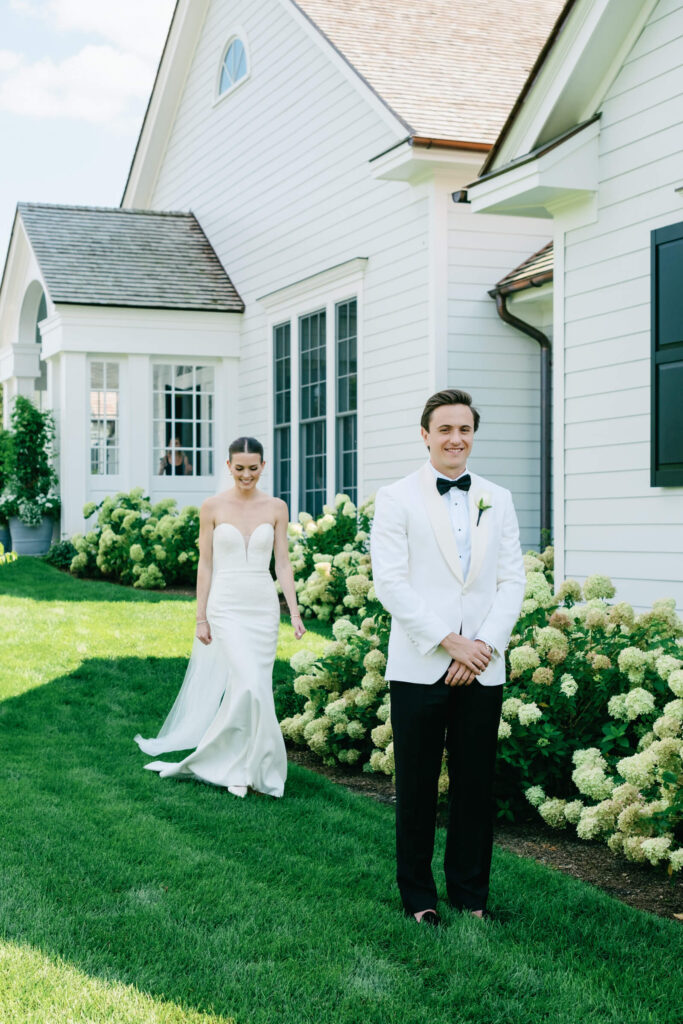 Bride walks toward groom for first look outside white country club building at Bedford Golf & Tennis Club