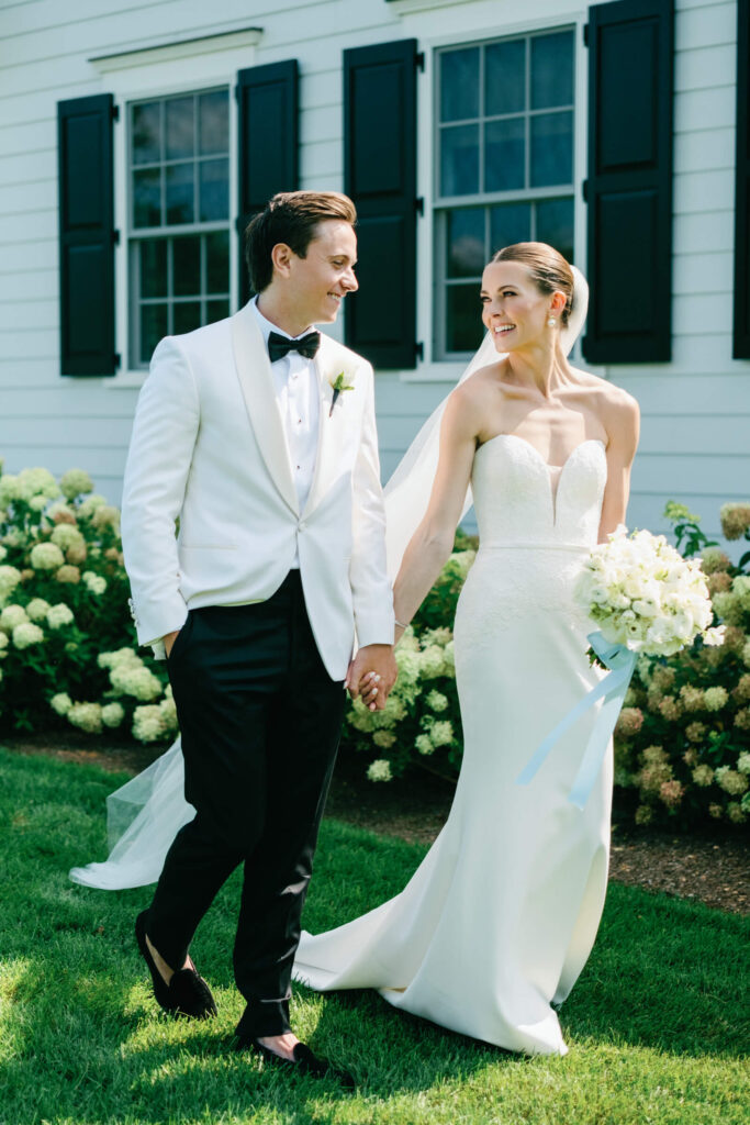 Bride and groom walk hand-in-hand after first look, smiling at each other in lush summer garden