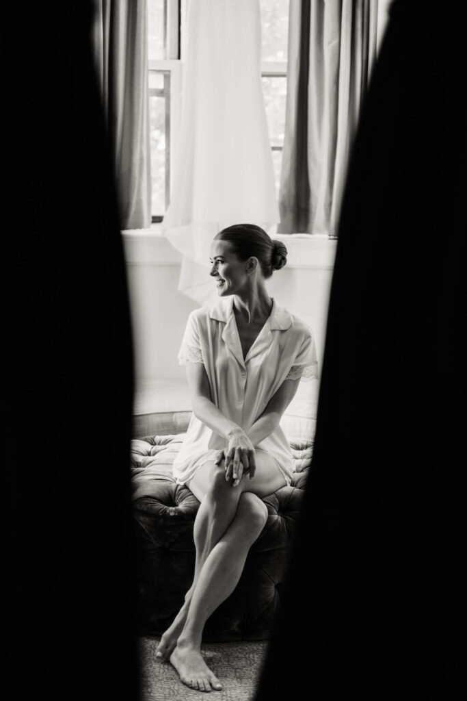 Black and white portrait of the bride in a robe, smiling and seated in the Castle Hotel & Spa suite with her wedding dress softly hanging behind her