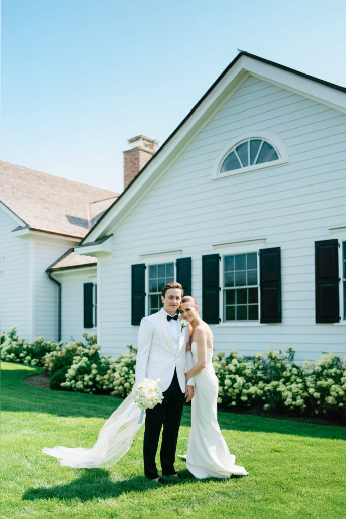 Elegant couple poses in front of the white house with black shutters and hydrangea bushes at New York Country Club