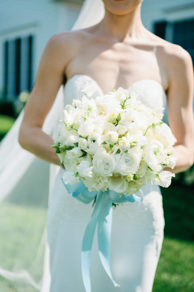 Close-up of the bride holding a lush bouquet of white ranunculus and roses tied with a soft blue ribbon