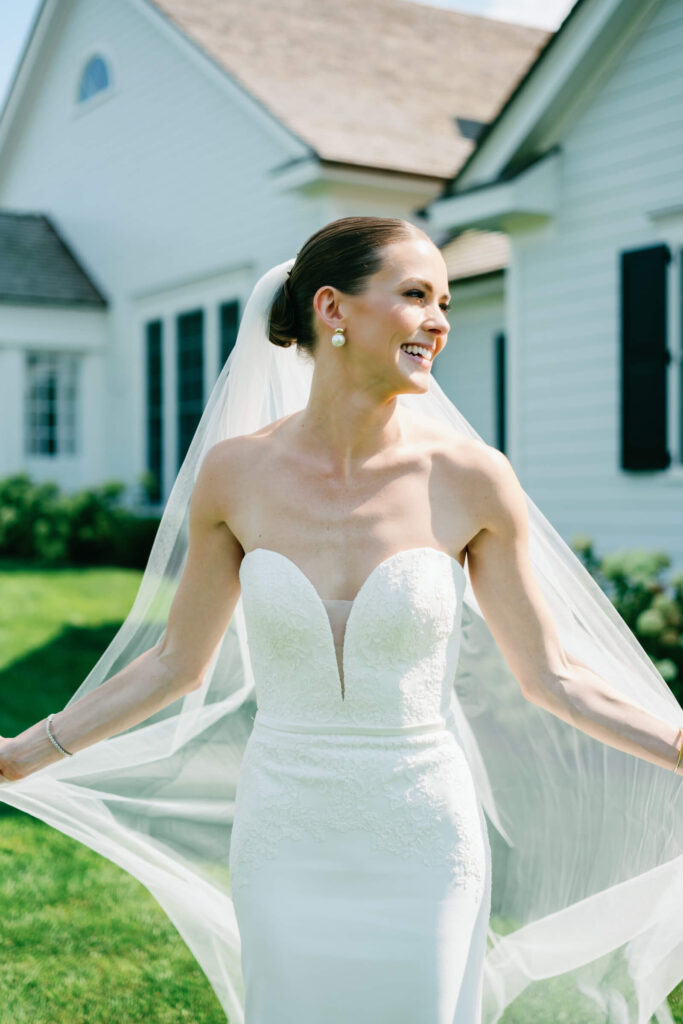 Radiant bride smiles and twirls, holding her flowing veil outside the New York Country Club estate
