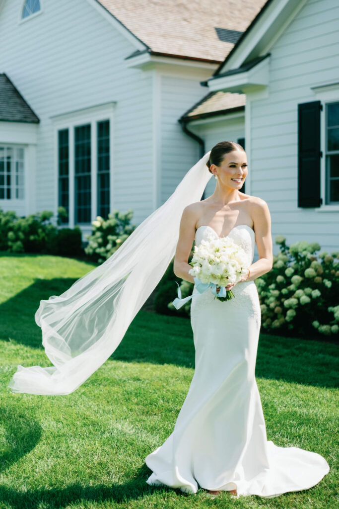 Bride stands gracefully with her bouquet and veil flowing in the breeze beside blooming hydrangeas
