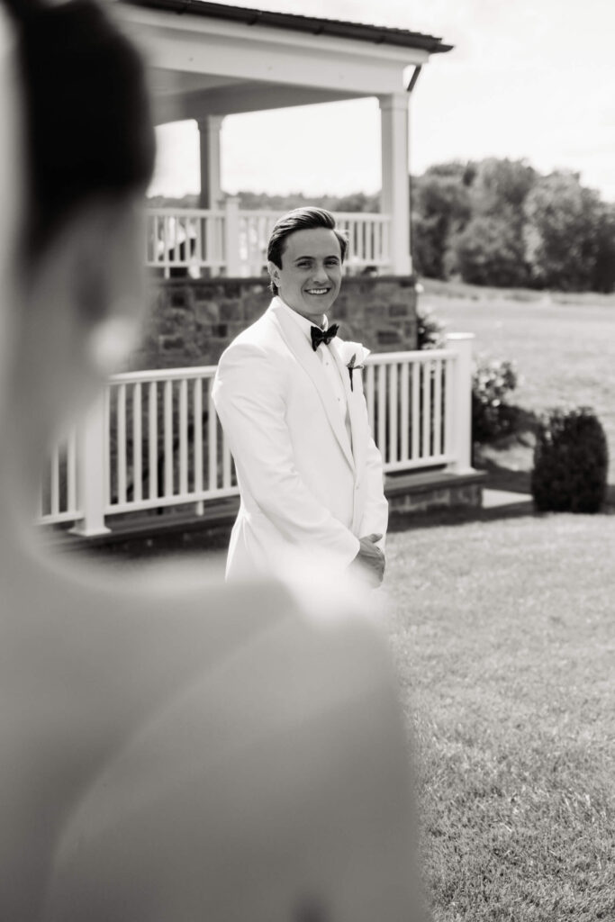Groom gazes lovingly at the bride from a distance in a black and white portrait outside the venue
