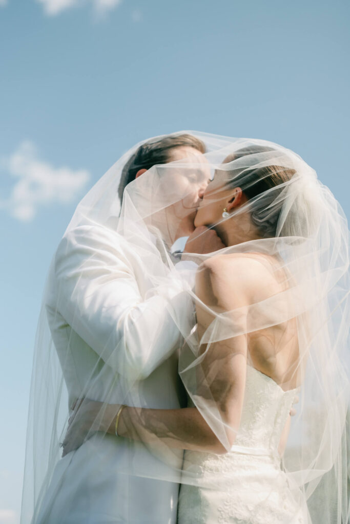 Bride and groom share a soft, ethereal kiss under the bride’s veil with a bright blue sky behind them