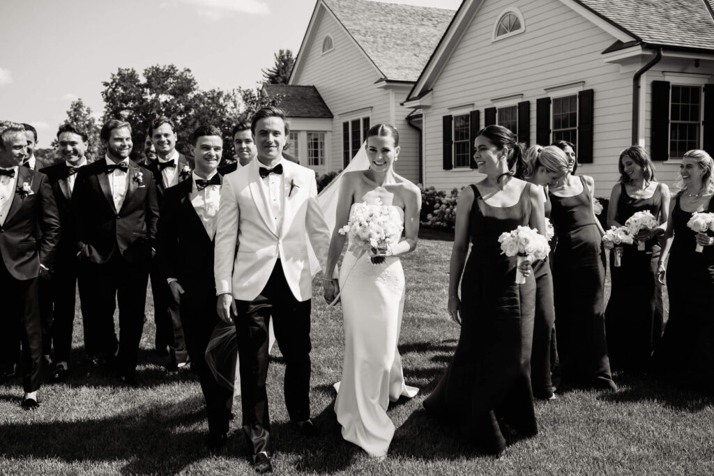 Wedding party walks together across the lawn, the groom in white and groomsmen in black tuxedos