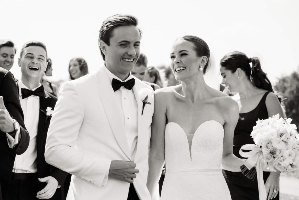 Candid moment of bride and groom smiling and walking hand-in-hand surrounded by joyful friends