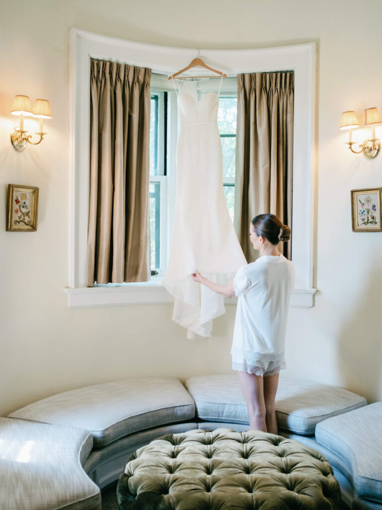 Bride reaching toward her gown hanging in a curved window nook at Castle Hotel & Spa during bridal prep on the morning of her New York country club wedding
