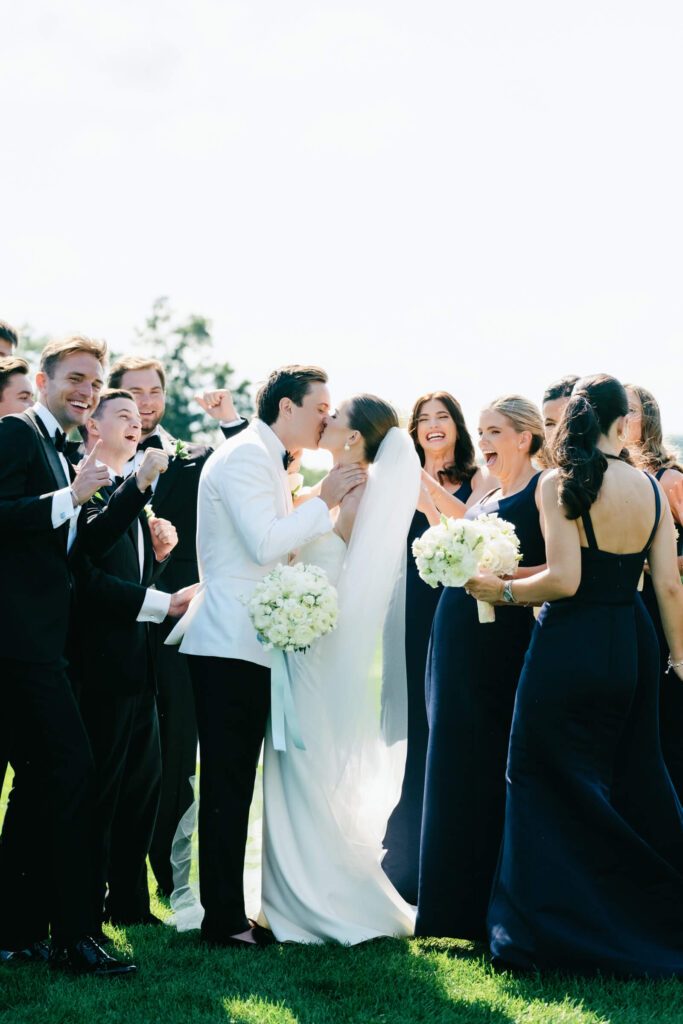 Bride and groom kiss surrounded by cheering wedding party during outdoor portraits at New York Country Club wedding
