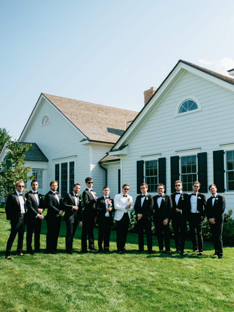 Groom and groomsmen in black tuxedos pose confidently outside white estate house at New York Country Club