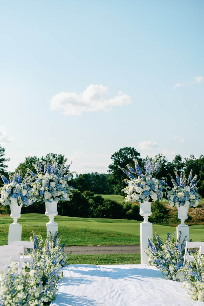 Lush blue and white floral arrangements in white urns frame the ceremony altar with sweeping golf course views in Bedford, NY