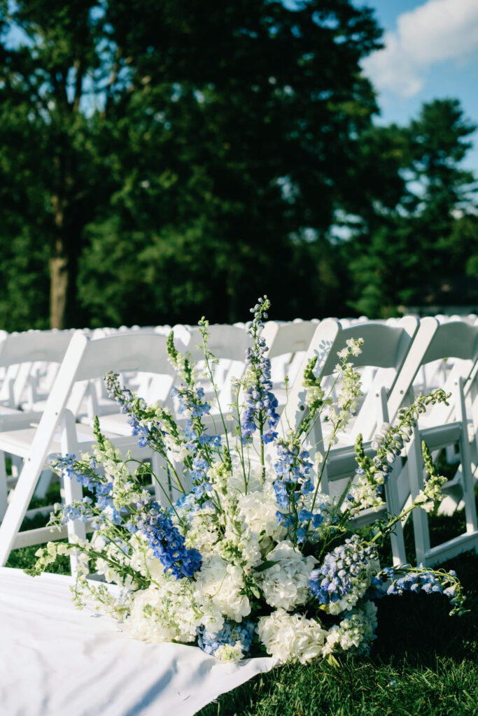 Ground-level view of romantic aisle florals with white and blue delphinium and hydrangea lining white guest chairs