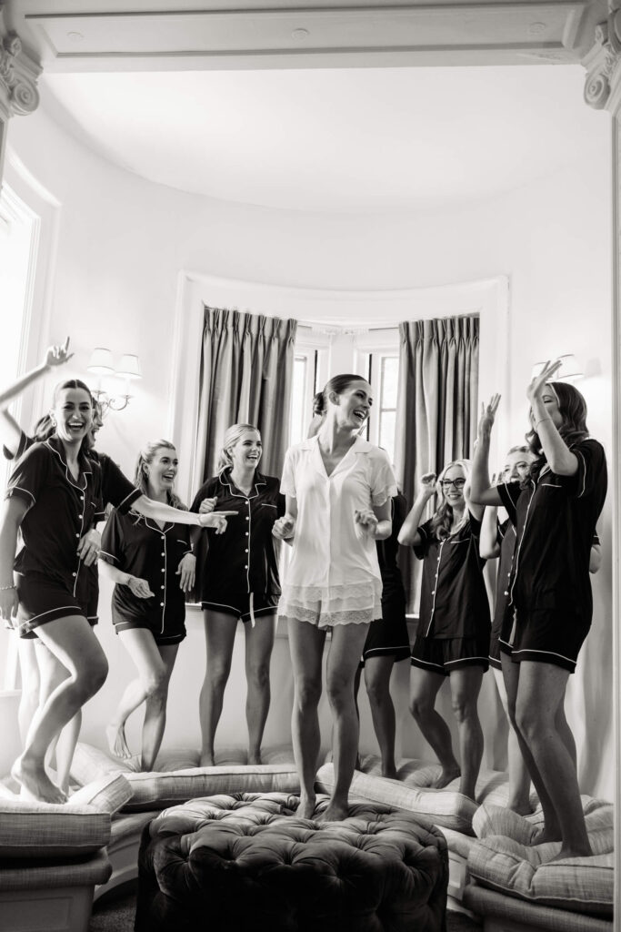 Black and white candid of the bride laughing with bridesmaids in matching navy pajamas, joyfully gathered in the bridal suite