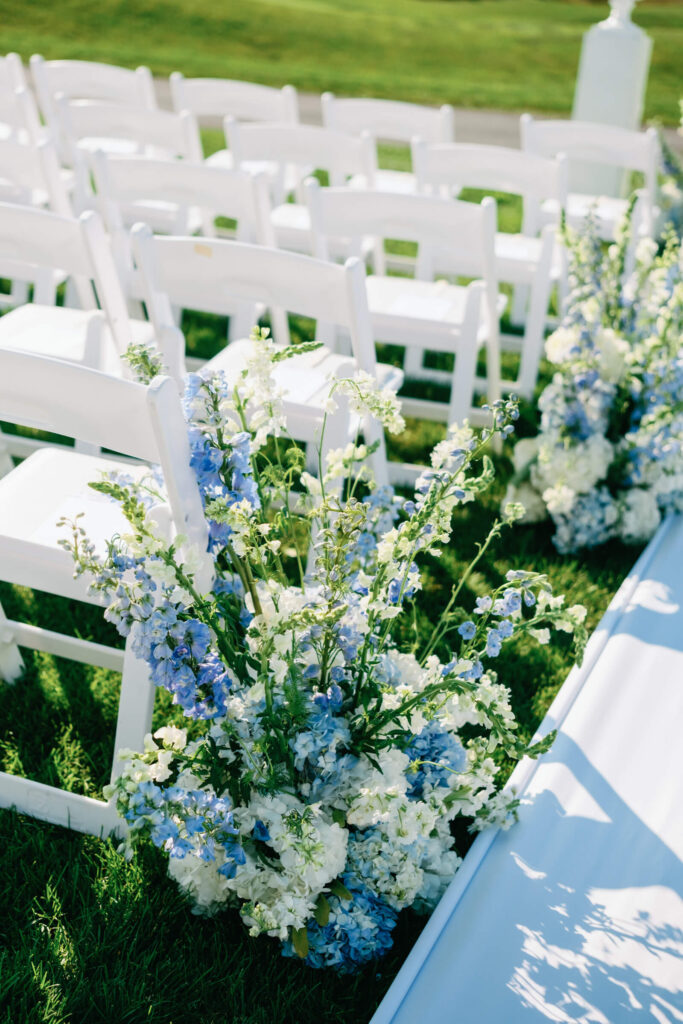 Close-up of white ceremony chairs flanked by lush floral arrangements in shades of blue and white lining the aisle at a New York Country Club wedding.
