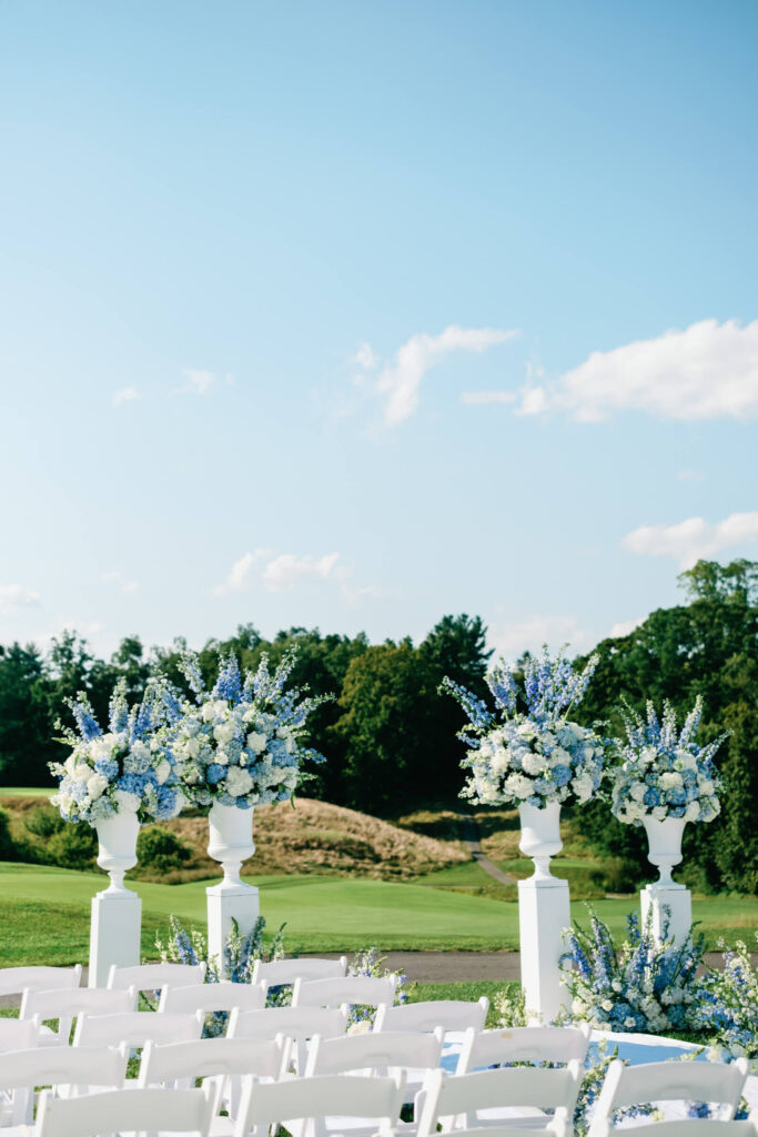 Blue and white flower arrangements on tall white pillars frame the ceremony aisle, with rolling green golf course hills in the background.