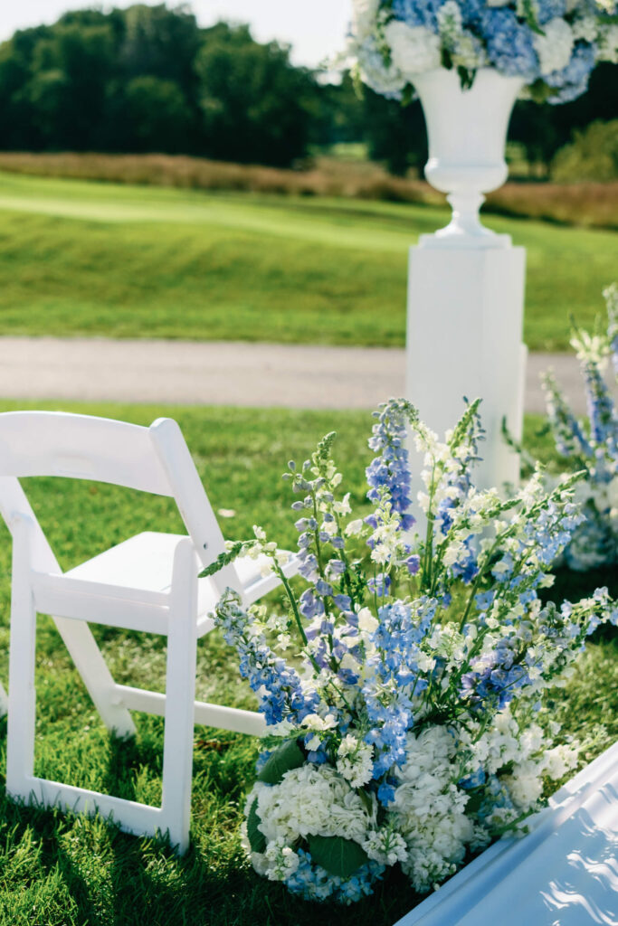 Lush floral installation with white hydrangeas and blue delphiniums next to a white folding chair at the ceremony site.