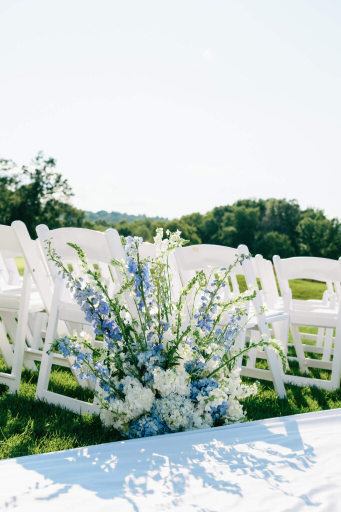 Romantic aisle floral arrangement in soft blue and white blooms placed beside a row of white guest chairs overlooking the green landscape.