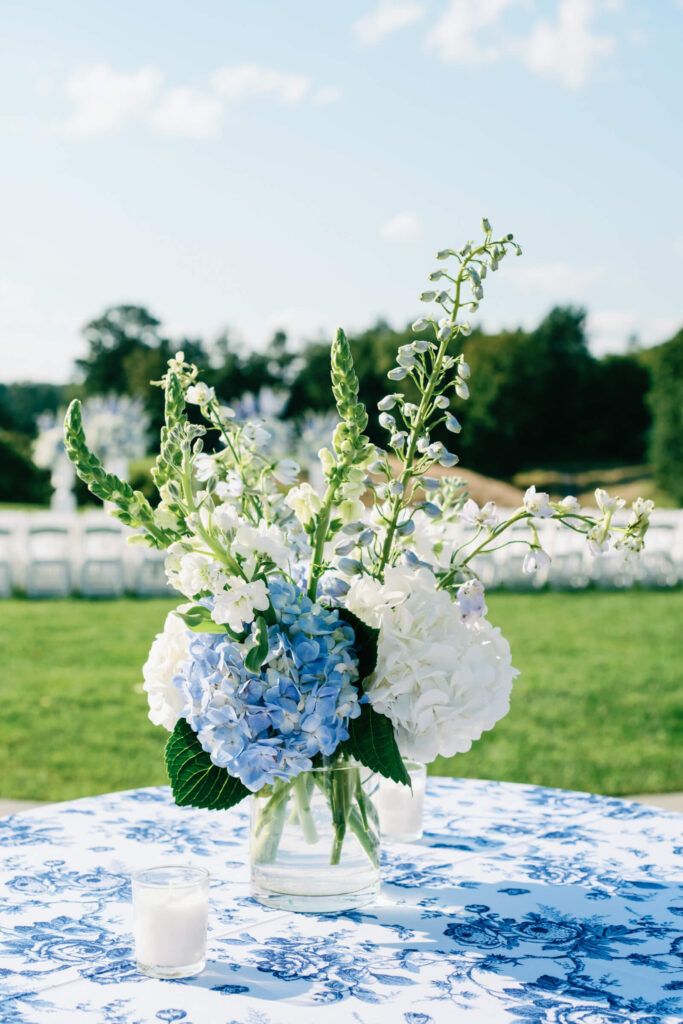 Close-up of a small centerpiece with blue hydrangeas, white stock, and snapdragons in a glass vase on a patterned cocktail table.
