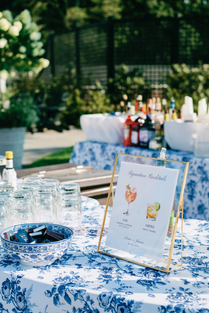 Signature cocktails display on a floral-patterned bar table, featuring framed signage for his Aperol Spritz and her spicy margarita, with a bowl of matchbooks.