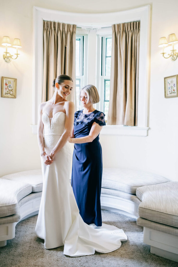 Bride smiling as her mother helps fasten the buttons on her classic strapless wedding gown in the Castle Parlor Suite