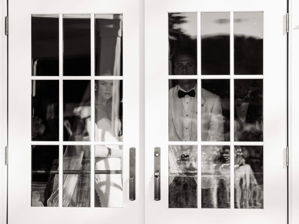 Artistic black-and-white portrait of bride and groom standing behind French doors, captured through the glass.