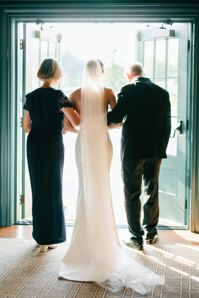 Bride standing between her parents, framed in the doorway just before walking down the aisle, with sunlight streaming in.