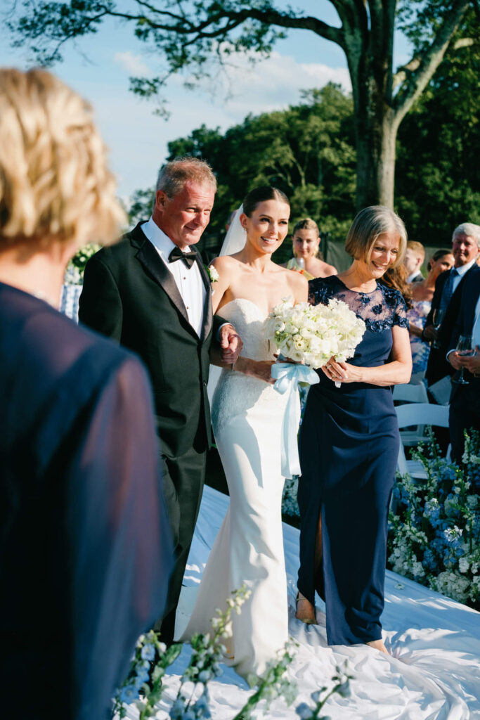 Bride walking down the outdoor ceremony aisle arm-in-arm with both parents, smiling as guests look on.