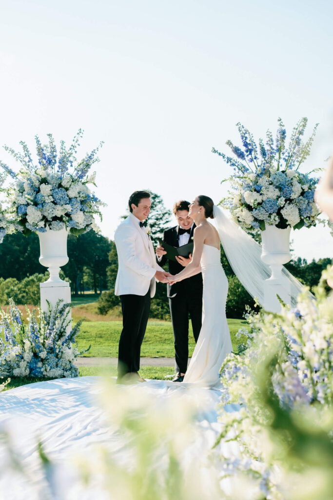 Couple holding hands and laughing at the altar during their outdoor ceremony surrounded by large floral arrangements.