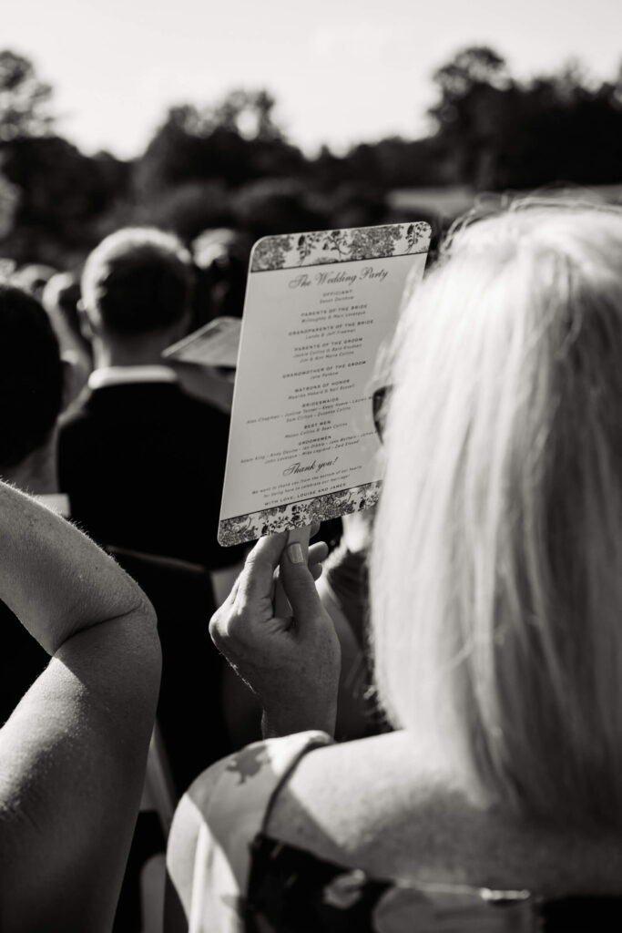 Wedding guest holding a printed program fan listing the bridal party, captured from behind during the ceremony.