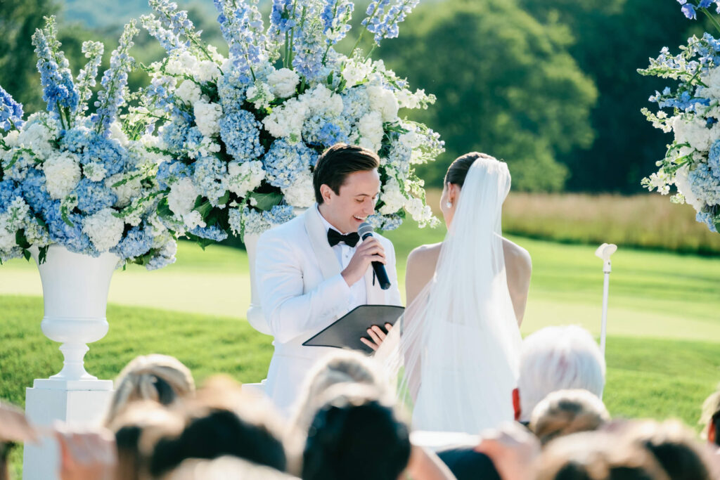 Groom reading vows with a microphone at the altar, with lush white and blue florals behind him.