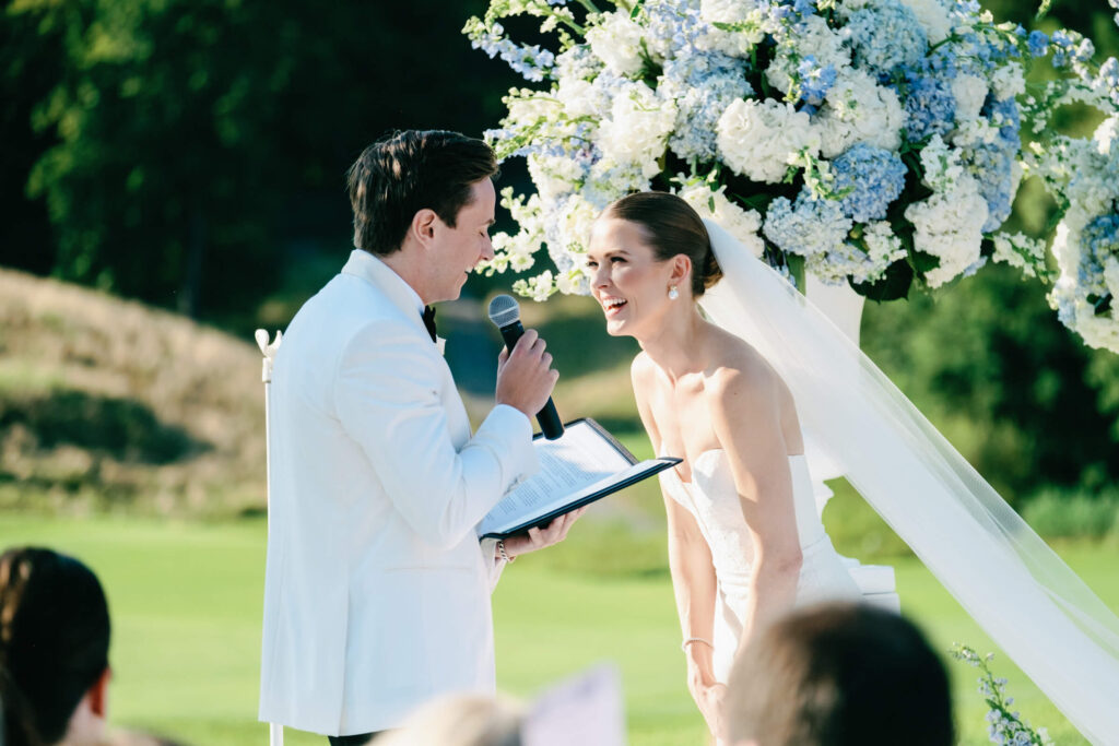 Bride smiling joyfully as the groom reads his vows, with elegant white and blue ceremony flowers in the background.