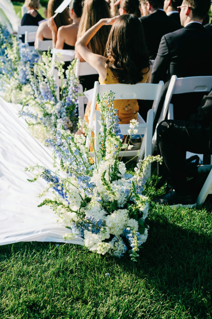 Guests seated along a flower-lined aisle at an outdoor ceremony, with white and blue floral arrangements in the foreground.