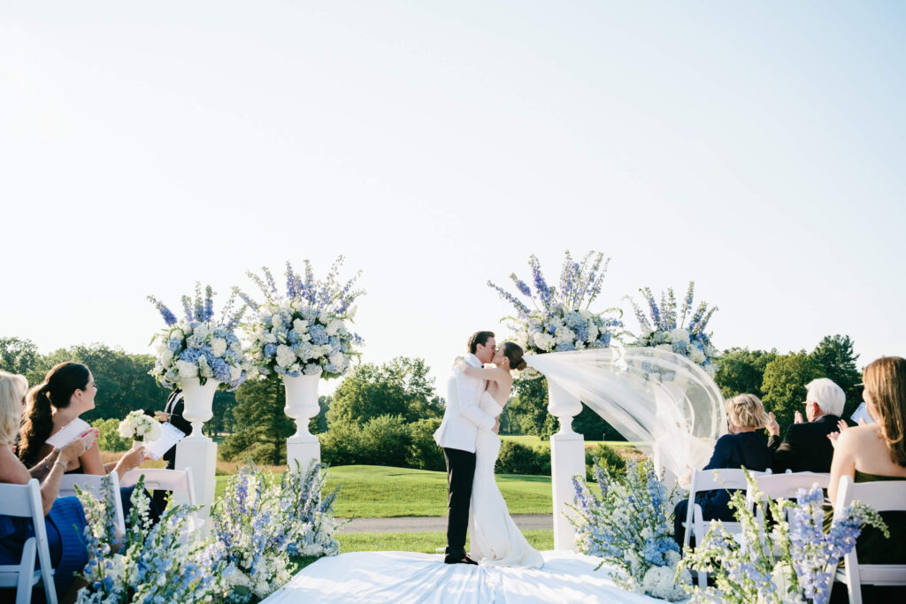 Bride and groom share their first kiss at the altar, surrounded by towering blue and white floral arrangements on the golf course at New York Country Club