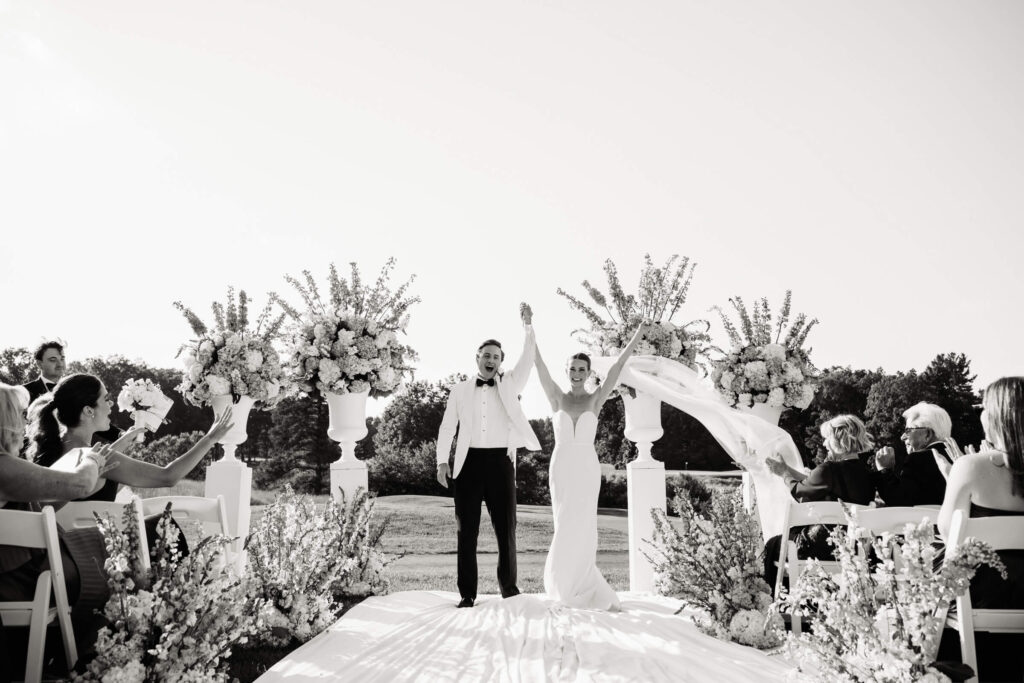 Newlyweds raise their hands in joy just after saying "I do," with the bride’s veil floating in the breeze and guests cheering them on