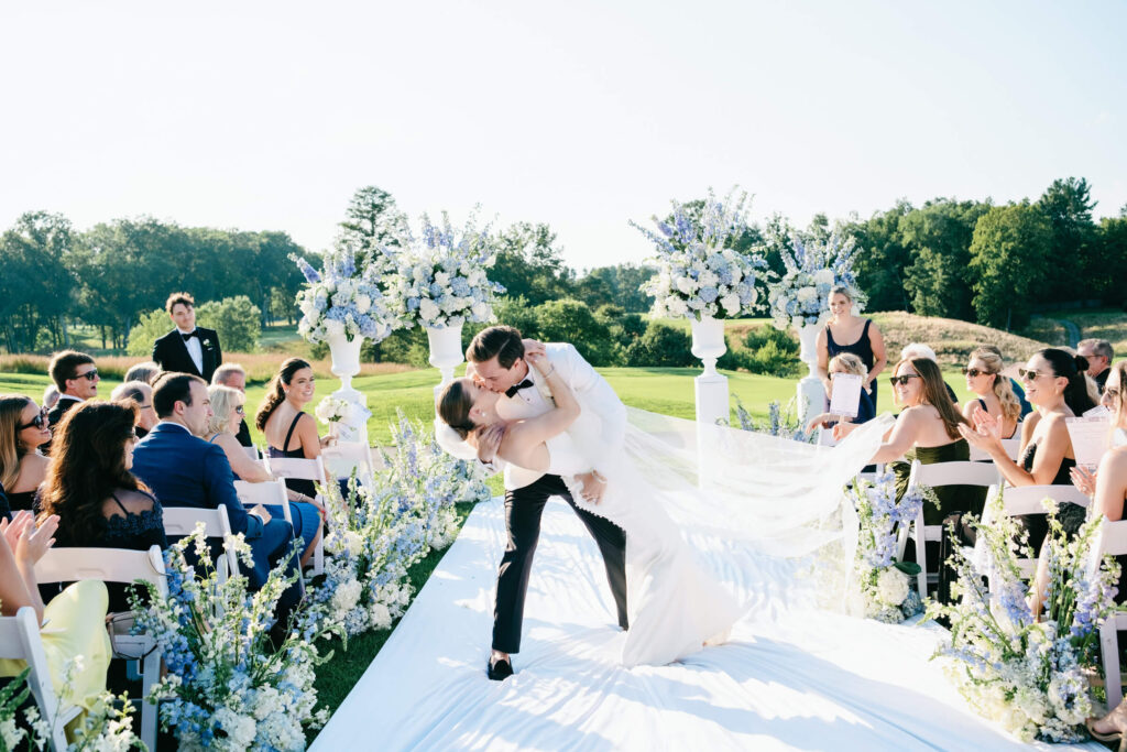 Groom dips his bride into a celebratory kiss as guests smile and applaud along a floral-lined ceremony aisle