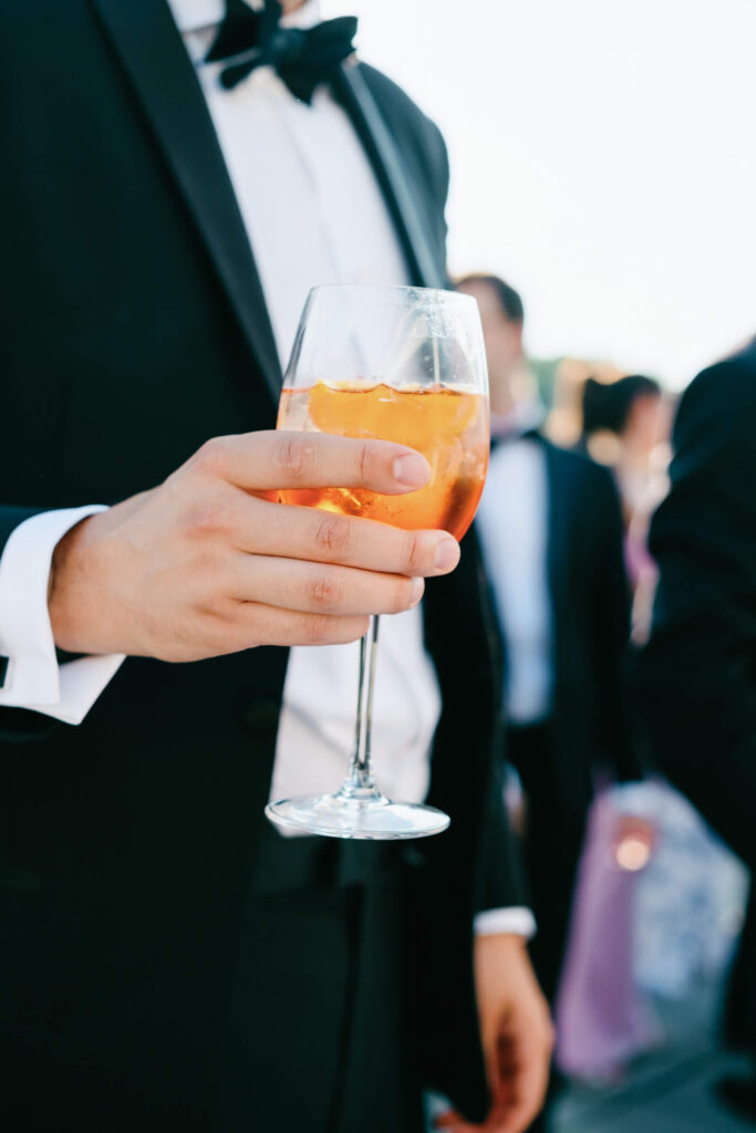 Close-up of a groom in black tie holding an Aperol Spritz cocktail during cocktail hour