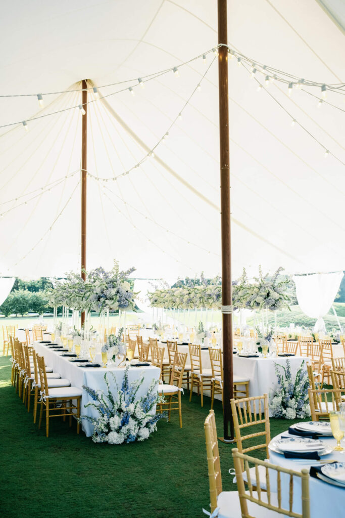 View inside the sailcloth reception tent, showcasing long banquet tables with lush floral arrangements and gold Chiavari chairs