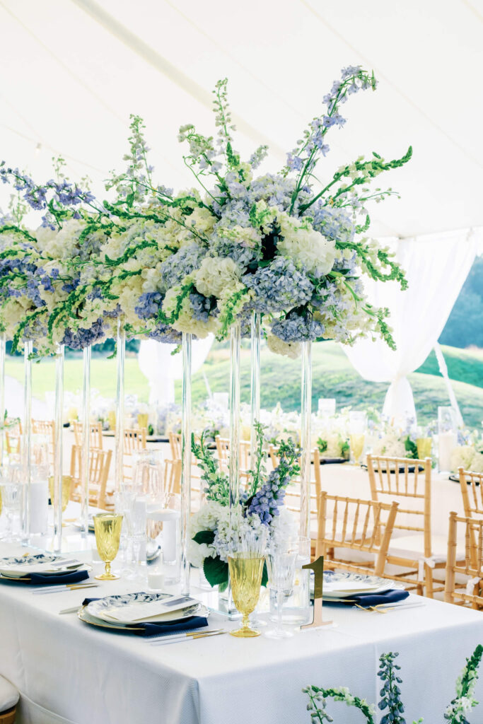 Tall floral centerpiece of white hydrangeas, delphinium, and stock blooms in glass vases, set on a table with layered place settings and golden accents