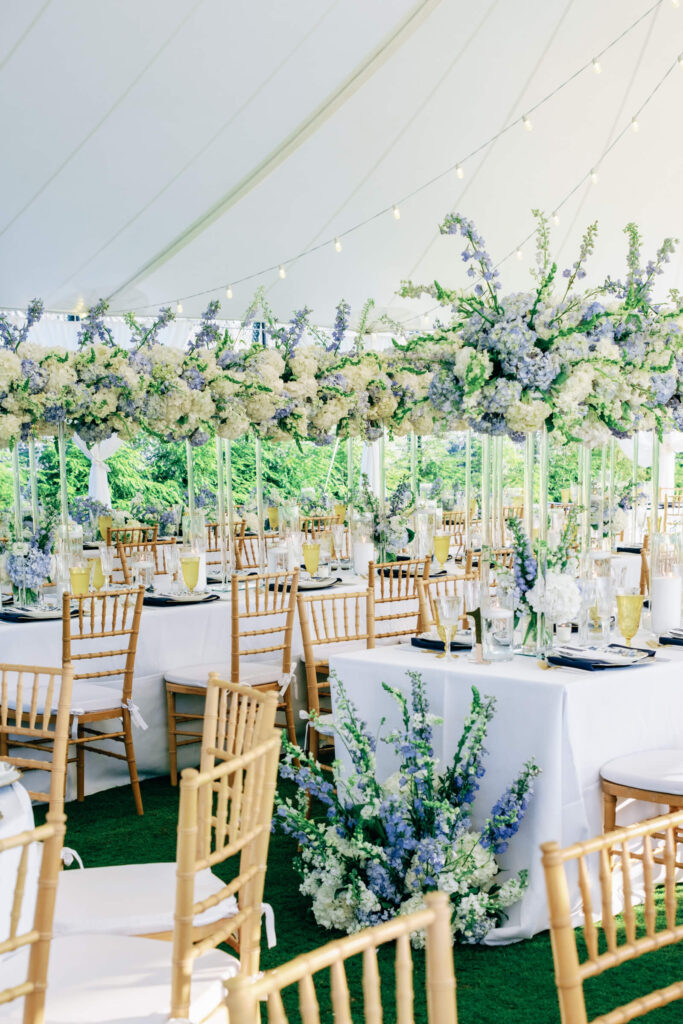 Long guest tables under a white sailcloth tent, elegantly adorned with tall centerpieces of white and blue florals, surrounded by gold Chiavari chairs.