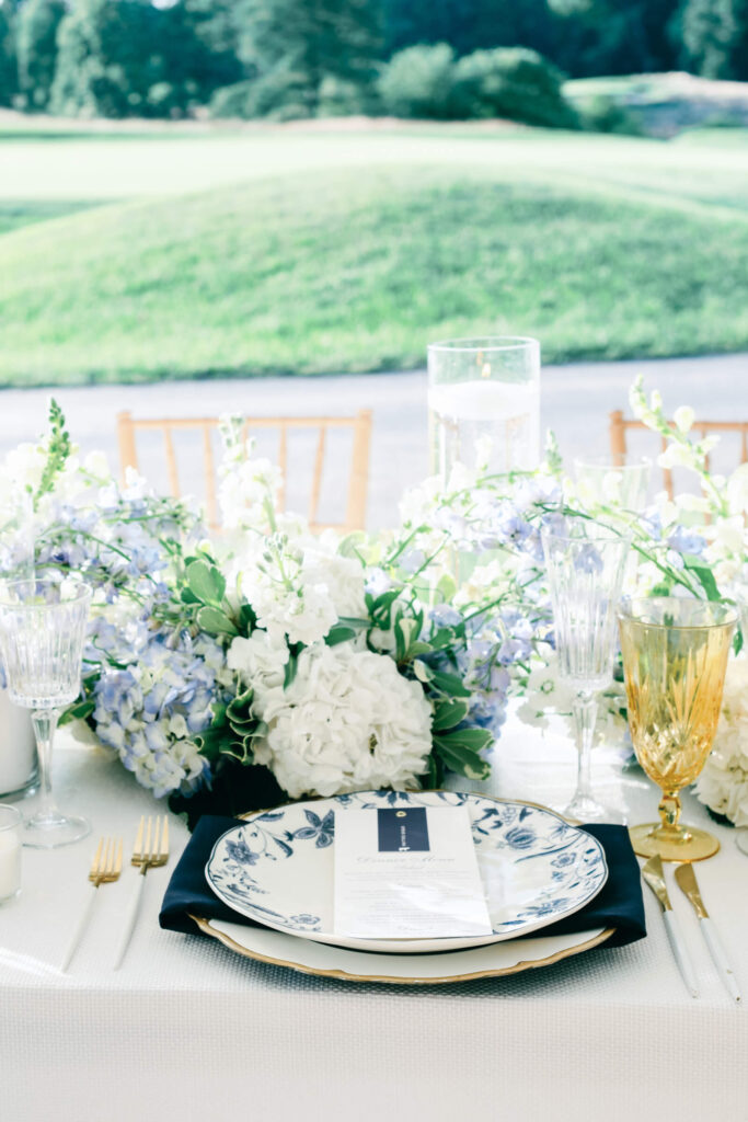 Reception table setting with layered blue and white china, golden flatware, and delicate glassware, framed by a lush low floral arrangement.