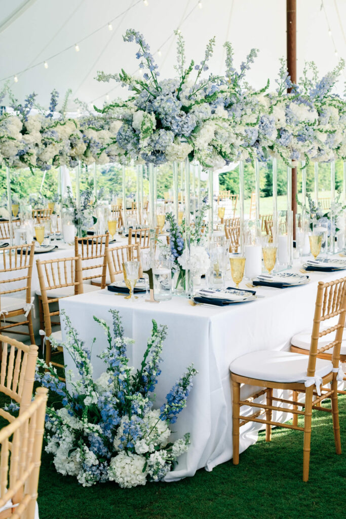 Side view of a beautifully set reception table with draped white linens, golden chairs, and low blue-and-white floral centerpieces under flowing white drapery.