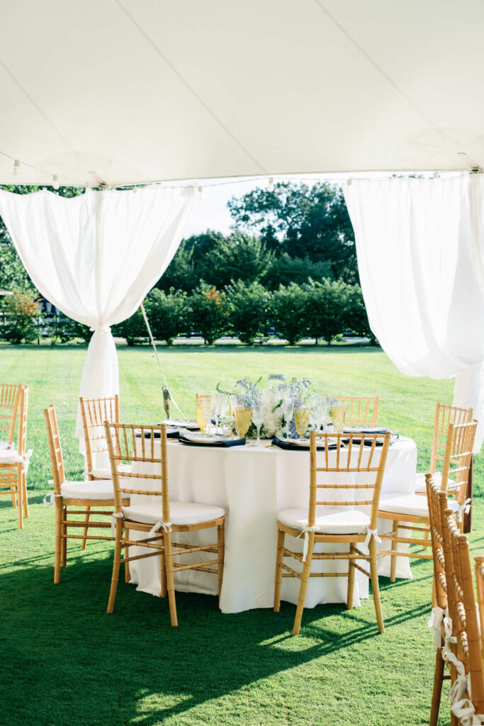 Round guest table tucked under a draped white tent opening, with garden views and fresh floral centerpieces echoing the wedding’s blue and white palette.